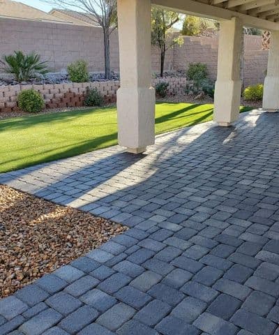Paved patio with pavers, landscaped yard, and shadows from pergola in sunny outdoor setting.