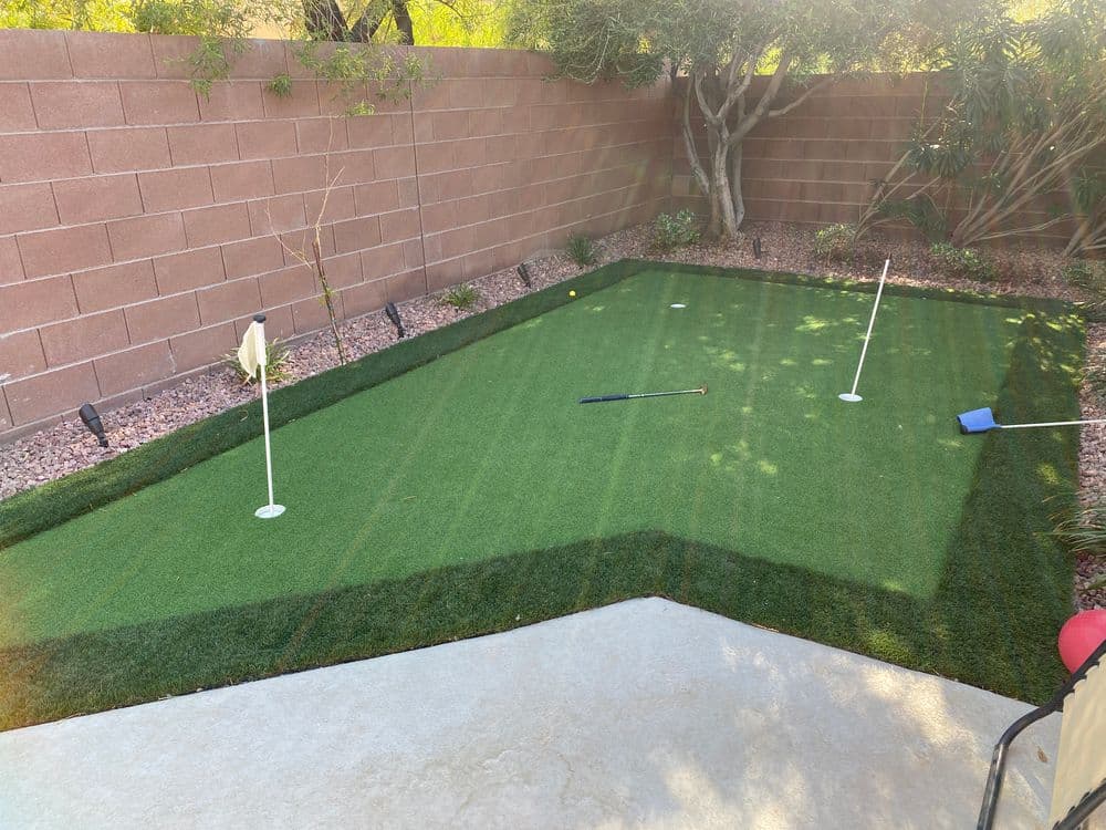 Backyard artificial putting green with flags, golf club, and lush landscaping. Perfect for golf practice.