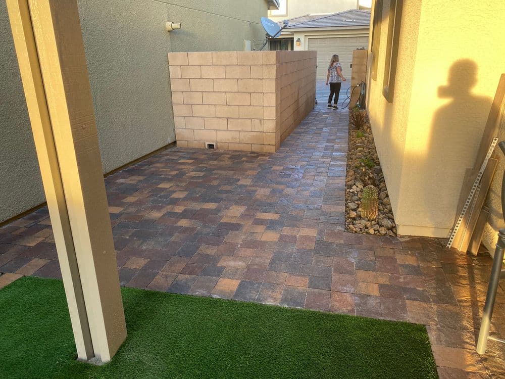 Paved backyard pathway with a person walking beside a wall and landscaped area.